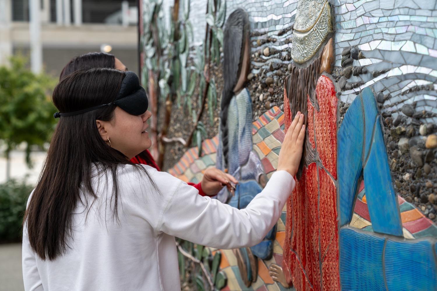 A young woman with long dark hair, wearing a white sweatshirt and with a padded mask covering her eyes, smiles as she runs her fingers along a sculpted mural depicting two people sitting on a blanket on a rocky beach.